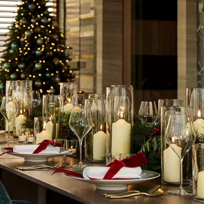 A festive dining table is set with white plates, napkins tied with red ribbon, gold cutlery, and tall glass candles. Greenery decorates the table. A decorated, lit Christmas tree is visible in the background.