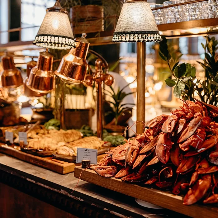 A buffet table features a large pile of cooked lobster claws and seafood under warm lights, surrounded by leafy garnishes and decorative greenery in an elegant restaurant setting.