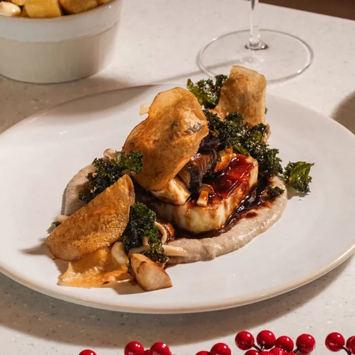A plated gourmet dish featuring a seared piece of fish atop a creamy puree, garnished with crispy potato chips, sautéed mushrooms, and fried kale, with a drizzle of dark sauce, served on a white plate.