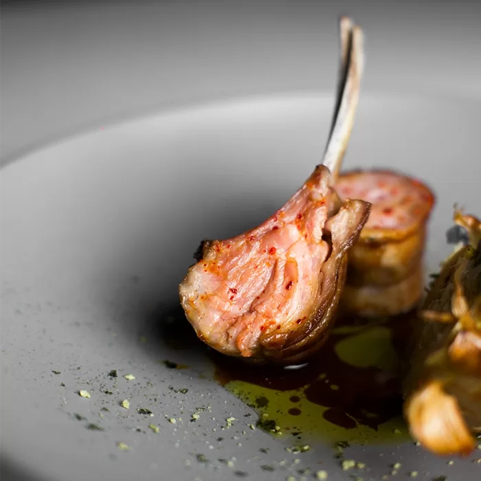 Close-up of a cooked lamb chop with a pink, juicy center on a white plate, garnished with herbs and sauce drizzled around it, creating a gourmet presentation.