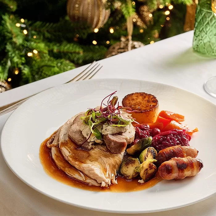 A festive plate of roast turkey with gravy, roast potatoes, Brussels sprouts, carrots, cranberry sauce, and pigs in blankets, garnished with microgreens. A decorated Christmas tree is in the background.