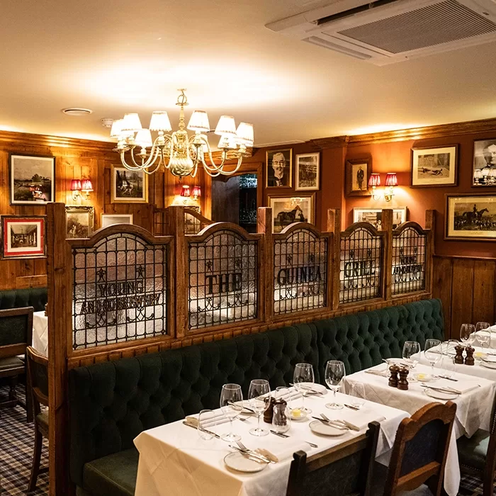 A cozy restaurant features white tablecloths, set tables, green tufted banquettes, wooden chairs, stained glass partitions, warm lighting, and framed pictures on wood-paneled walls, creating an inviting atmosphere.
