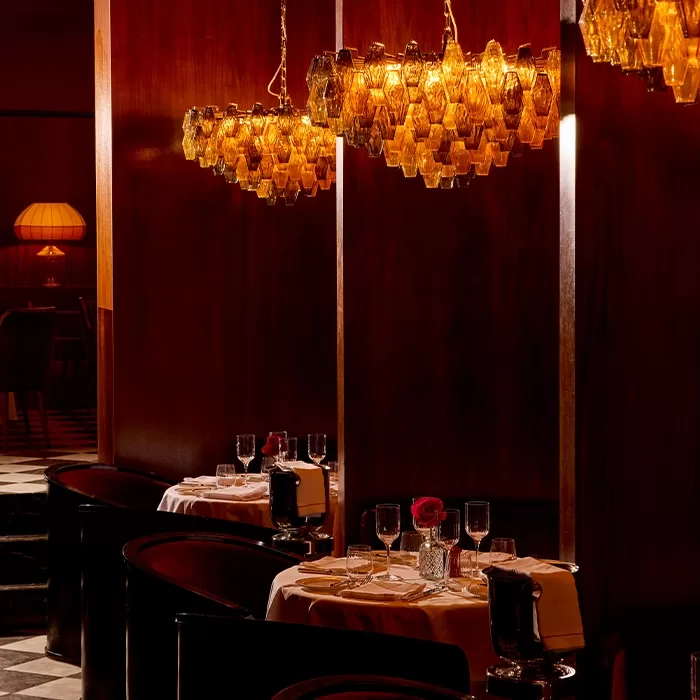 A dimly lit, elegant restaurant with dark wood walls, amber glass chandeliers, round tables set with white tablecloths, wine glasses, and a single red rose in a vase, creating an intimate atmosphere.