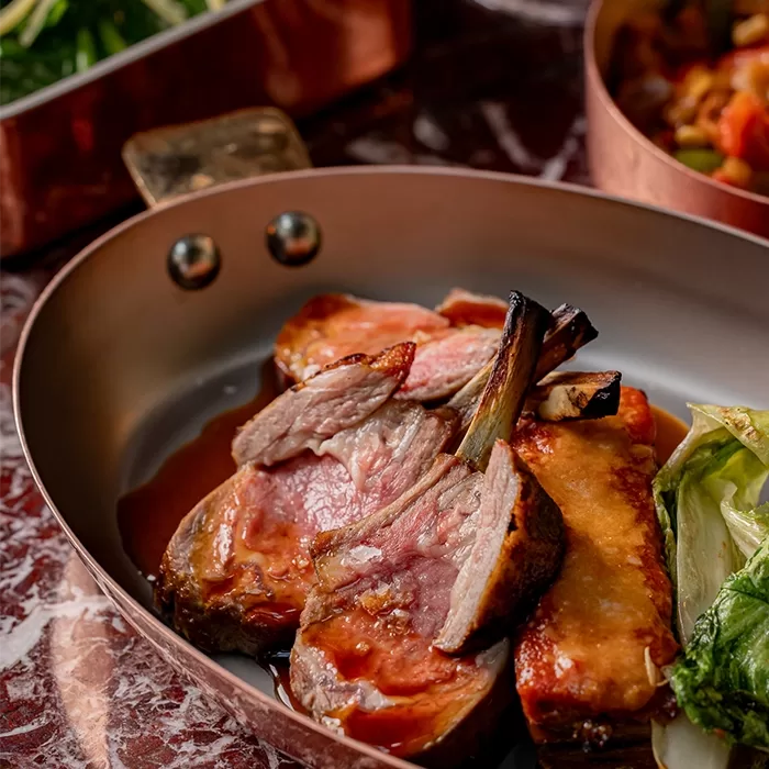 A copper pan filled with slices of medium-rare lamb chops, a thick piece of lamb with caramelized crust, and a portion of cooked green vegetables on a red marble surface.