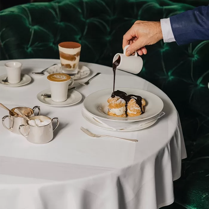 A person pours chocolate sauce over cream-filled pastries on a white plate, set on a round table with coffee drinks, a sugar bowl, and a green velvet couch in the background.
