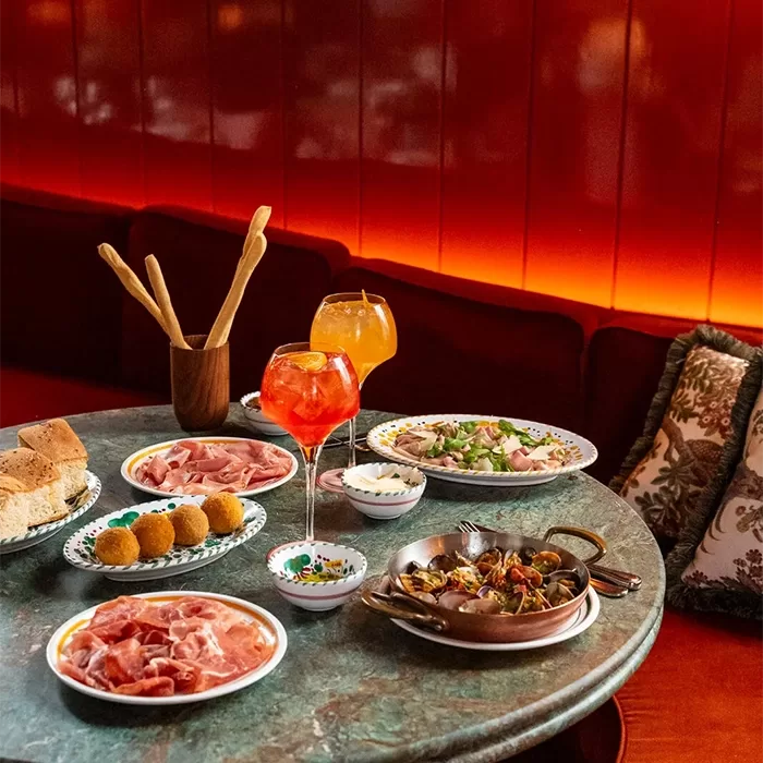 A round table set with Italian appetizers, including cured meats, breadsticks, croquettes, seafood, and cocktails, against a red cushioned booth with patterned pillows and warm ambient lighting.