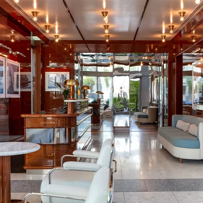 A stylish hotel lobby with glossy wood walls, marble floors, elegant white chairs, a marble table, modern sofas, gold lamps, artwork, and a mirrored entrance reflecting greenery outside.