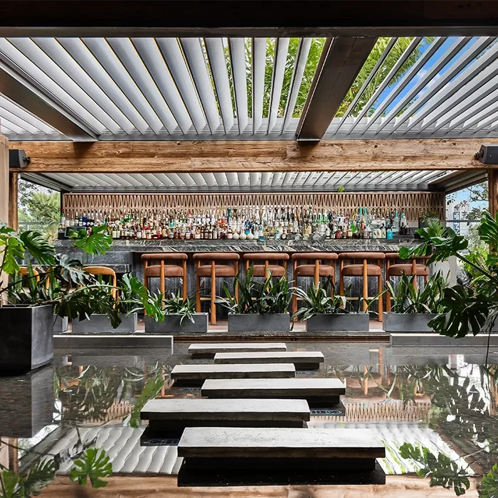 A modern outdoor bar with wooden stools, lush green plants, and a water feature with stone steps leading to the bar; overhead, a pergola with angled slats lets in natural light. Bottles line the back wall behind the bar.