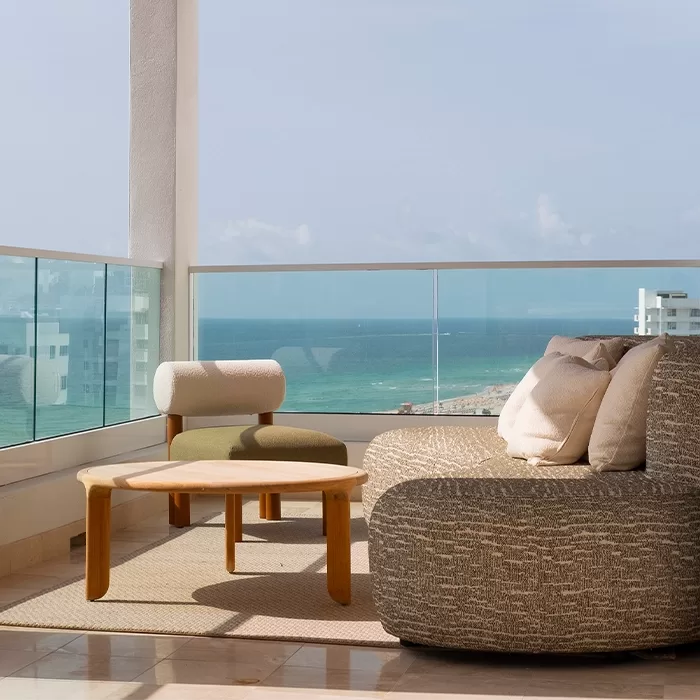 Modern balcony with a round cushioned chair, a wooden armchair, and a low wooden table on a rug, overlooking a clear blue ocean and sky through a glass railing.