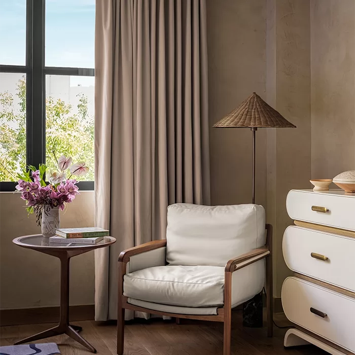 A cozy corner with a white cushioned chair, a round wooden side table holding a vase of flowers and a book, a tall floor lamp with a woven shade, and a cream dresser near a window with beige curtains.