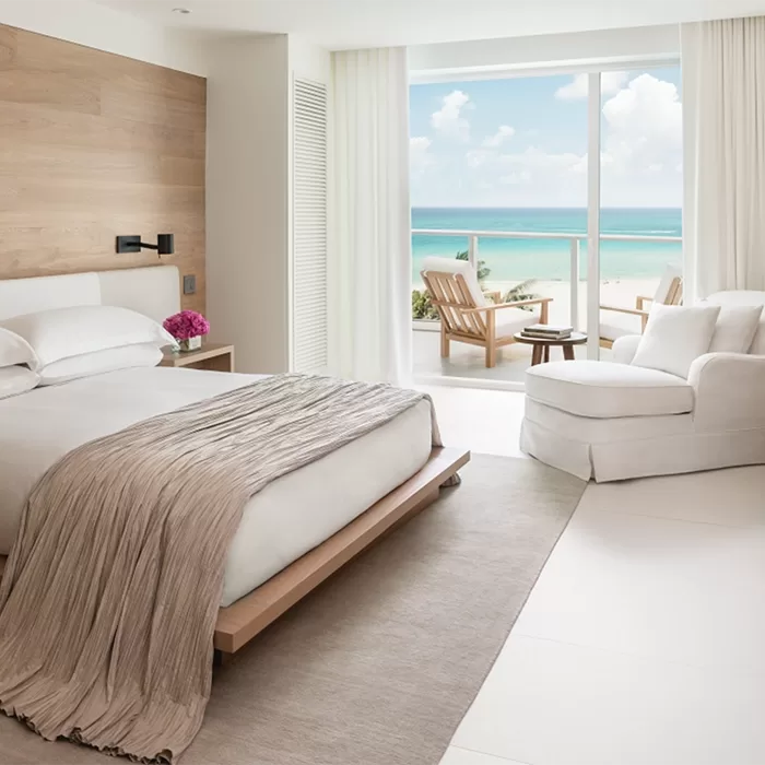 Modern hotel room with a large bed, light neutral decor, wooden accent wall, white armchair, and sliding glass doors opening to a balcony with chairs, overlooking a blue ocean under a partly cloudy sky.