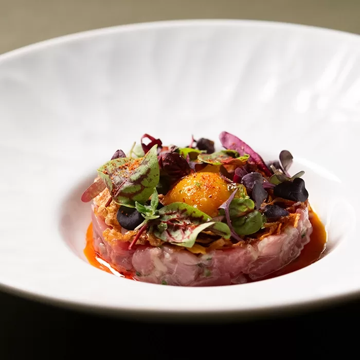 A gourmet dish featuring finely diced raw fish arranged in a circular shape, topped with a raw egg yolk, microgreens, and colorful edible leaves, served with a vibrant sauce on a white plate.