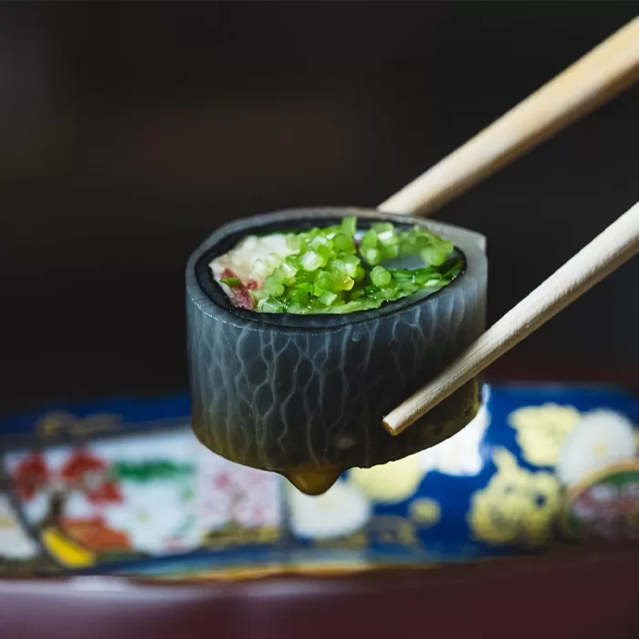 A close-up of chopsticks holding a piece of sushi wrapped in dark seaweed, filled with green vegetables, over a colorful, patterned dish.