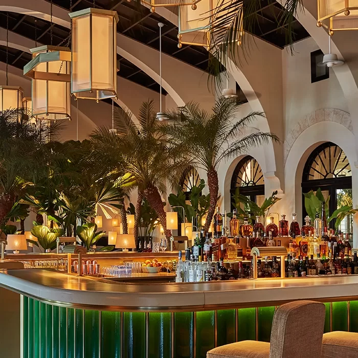 A stylish bar with a green tiled counter, surrounded by lush indoor palm trees and plants, warm lighting from large hanging lanterns, and arched windows in the background. Bottles and glasses are neatly arranged on the bar.