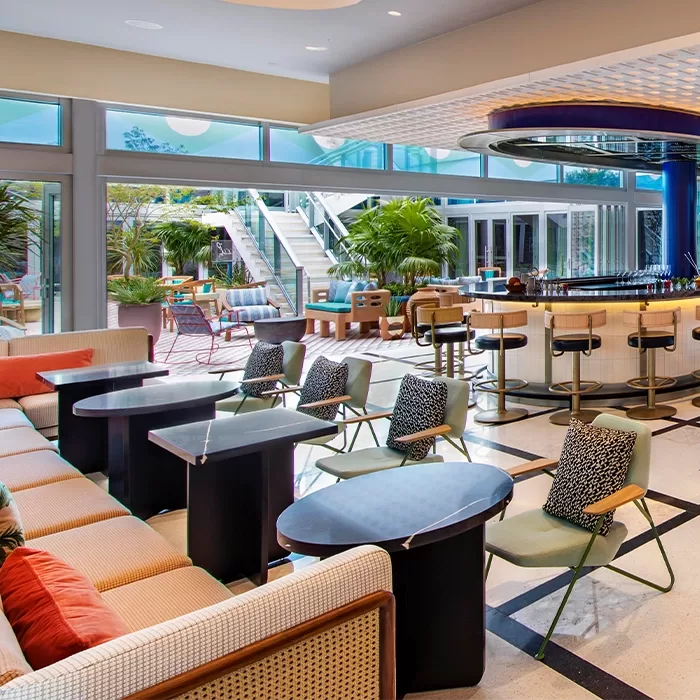 A modern, stylish hotel lounge with colorful seating, cushioned chairs, black tables, a curved bar with stools, large windows, potted plants, and outdoor patio access with steps leading to an upper terrace.