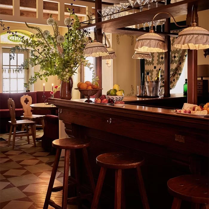 Cosy bar with wooden stools, a polished wooden counter, hanging glass lamps, fresh fruit bowls, a vase with tall flowers, and a softly lit, inviting dining area in the background.