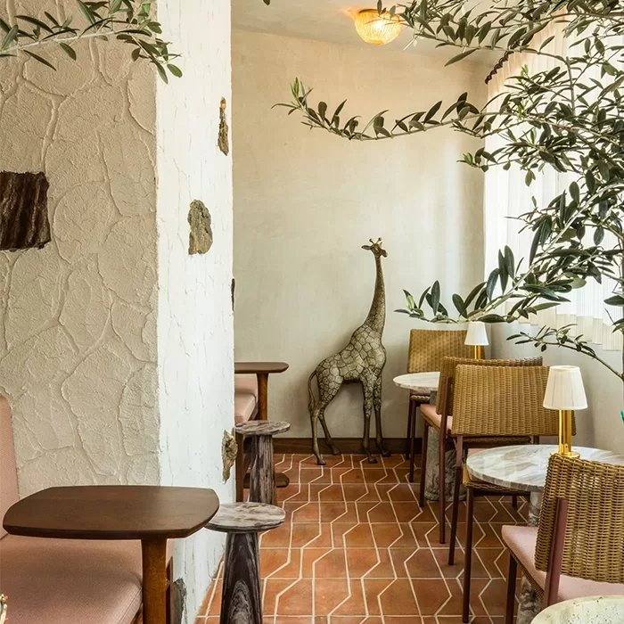 A cosy café corner with wooden tables and wicker chairs, decorated with potted plants, earthy textures, and a life-sized giraffe sculpture near the window. Sunlight filters in, creating a warm and inviting atmosphere.