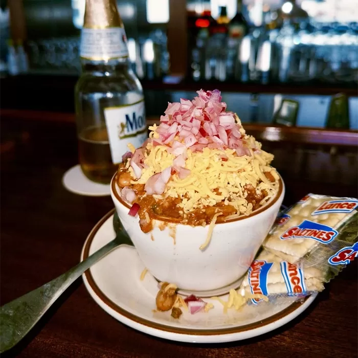 A bowl of chili topped with shredded cheese and chopped red onions sits on a plate with a spoon, a pack of Lance Saltines crackers, and a bottle of Modelo beer in the background on a bar counter.