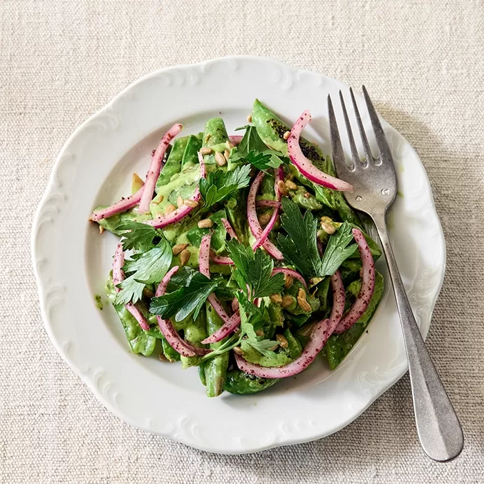 A white plate holds a fresh salad with snap peas, pickled red onions, parsley, and sunflower seeds, served with a fork on a light, textured tablecloth.