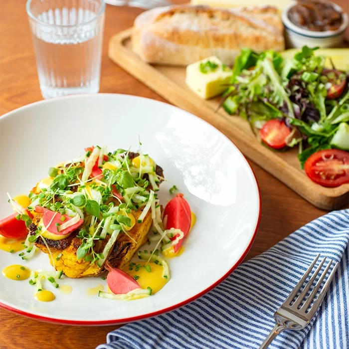 A plated gourmet meal with grilled toast topped with microgreens, radishes, and yellow sauce, beside a salad, cheese, bread, and a glass of water on a wooden table with a striped napkin and fork.