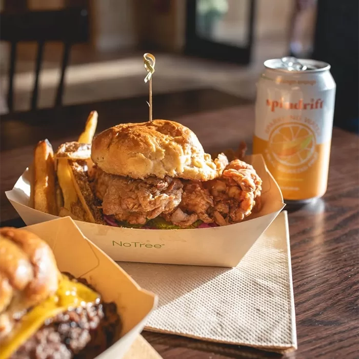 A crispy fried chicken sandwich with lettuce and tomato sits in a paper tray with golden fries, next to a can of Spindrift sparkling water on a wooden table. Sunlight streams in, illuminating the meal.