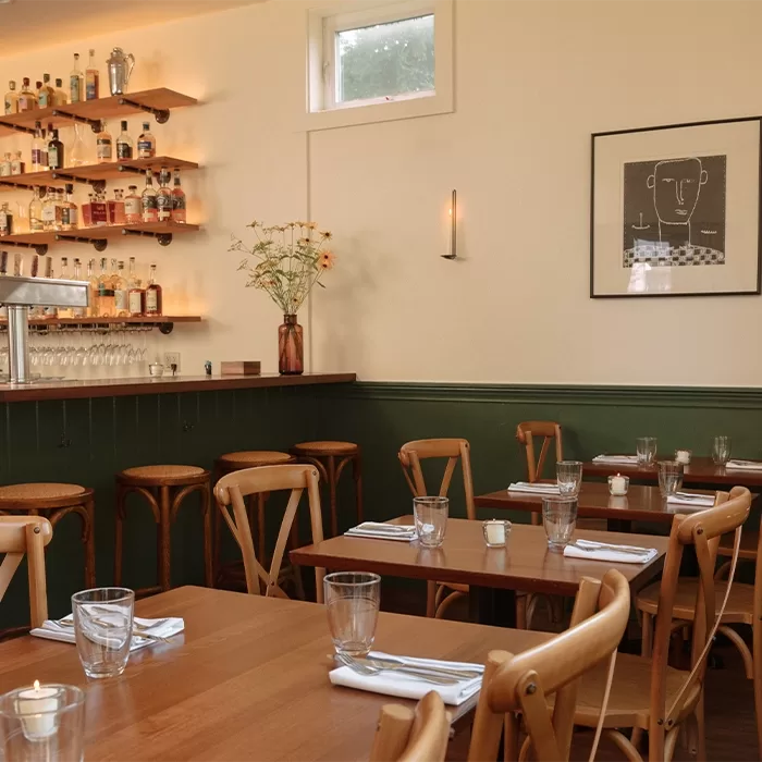 Cozy restaurant interior with wooden tables and chairs, neatly set with glasses and napkins. A small bar with shelves of bottles, a vase of flowers, and wall art add to the warm, inviting atmosphere.