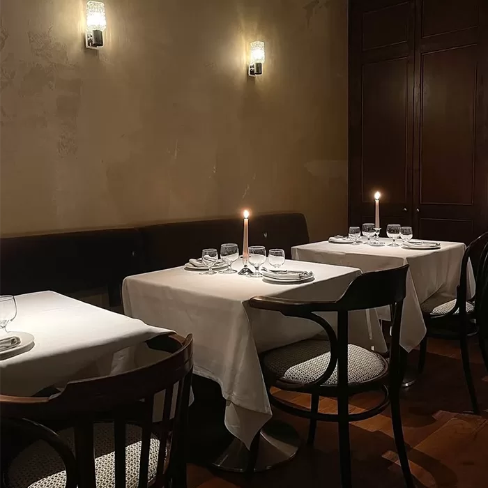 A dimly lit restaurant dining area with neatly set tables covered in white cloths, each featuring a lit candle, glassware, and napkins. Dark chairs and soft wall lighting create a cozy, intimate atmosphere.