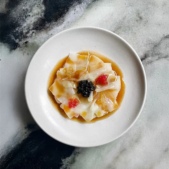 A white plate with thinly sliced raw fish arranged in a circle, topped with a dark center garnish, small red dollops, and drizzled sauce, set on a marbled surface.