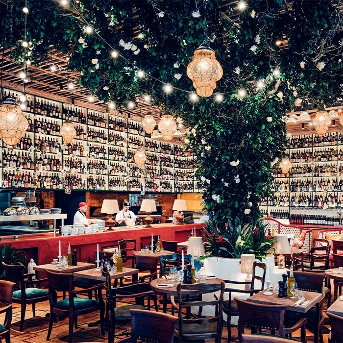 A stylish restaurant with walls lined with shelves full of wine bottles, lush greenery and string lights hanging from the ceiling, cozy wooden tables and chairs, and a bar area where chefs are preparing food.