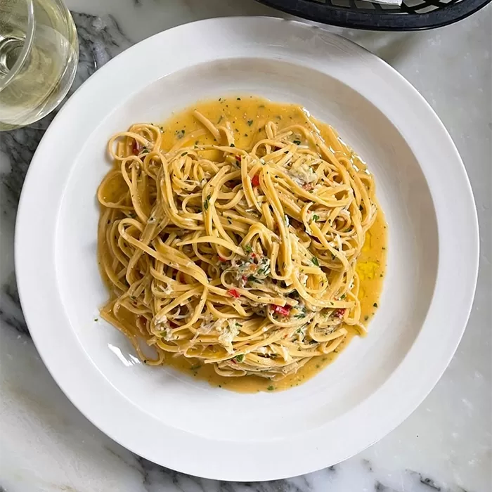 A white plate of spaghetti coated in a creamy yellow sauce with herbs and red pepper flakes, served on a marble table next to a glass of white wine.