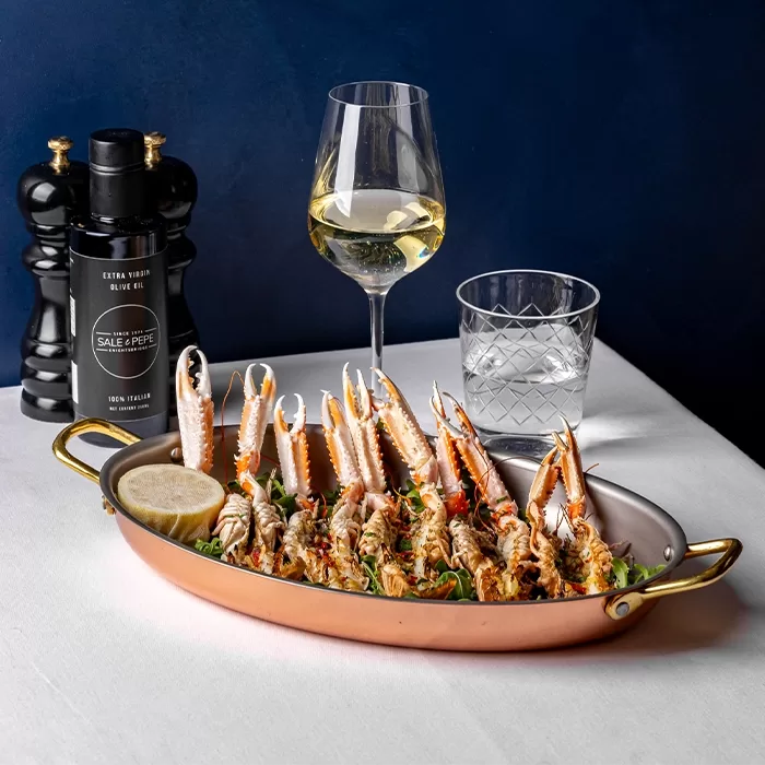 A dish of cooked langoustines garnished with herbs and a lemon slice is served in a copper pan. Nearby are a glass of white wine, a glass of water, and black pepper and olive oil bottles on a white tablecloth.