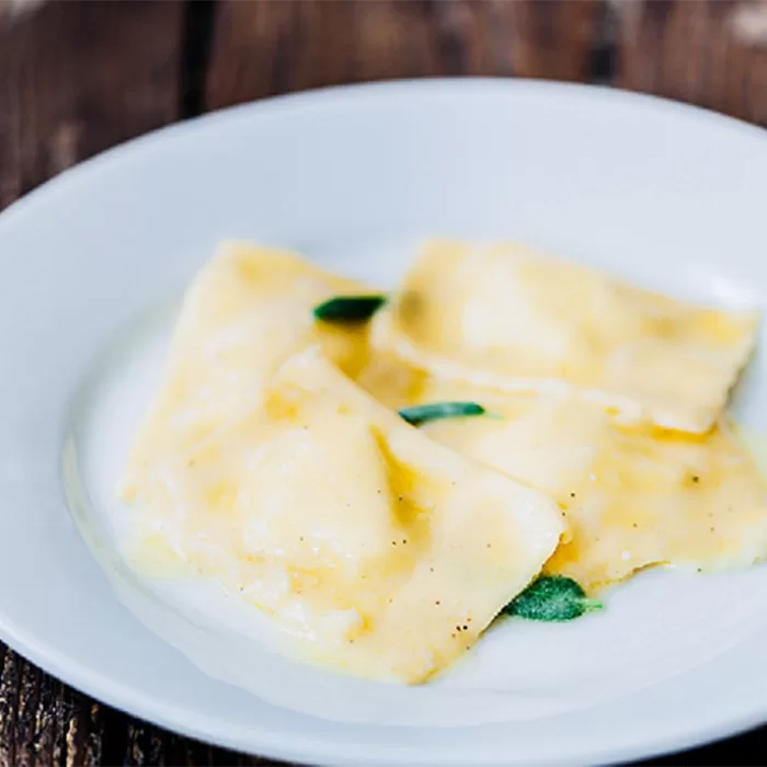 Three pieces of ravioli with a light butter sauce and sage leaves are served on a white plate, set on a rustic wooden table.
