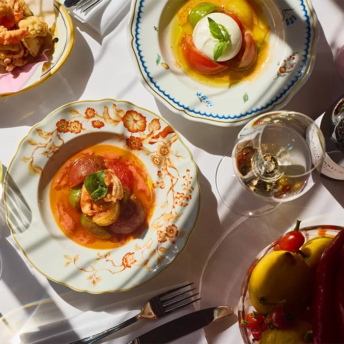 Plates of colorful tomato and mozzarella salads, garnished with basil, are set on a white tablecloth alongside wine, cutlery, and fresh vegetables under bright natural light.