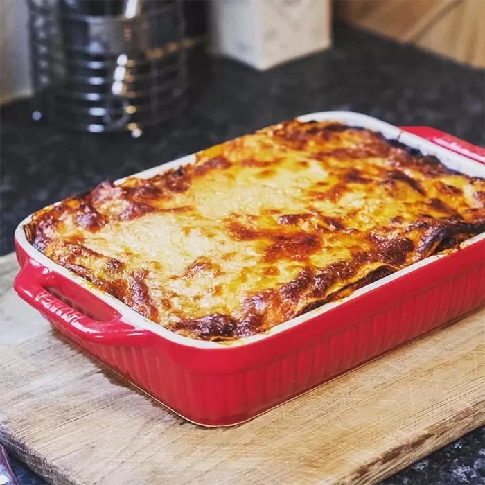 A freshly baked lasagna with a golden, melted cheese topping sits in a red rectangular baking dish on a wooden cutting board in a kitchen.