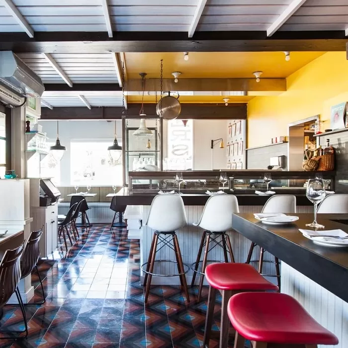 Bright, modern restaurant interior with colorful geometric tile floor, white and red chairs, a black counter, hanging lights, and a yellow accent wall behind the bar area. Tables are set with glasses and cutlery.