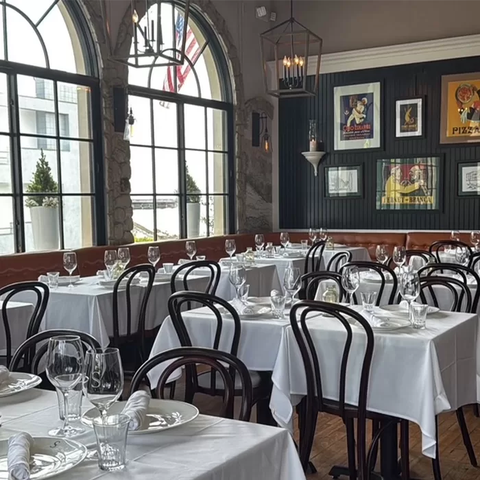 A bright, elegant restaurant with white tablecloths, dark wooden chairs, neatly set tables, and large arched windows. Framed art and posters decorate the dark-paneled wall.