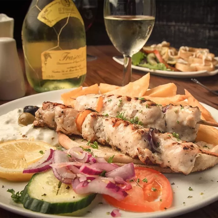 A plate of grilled chicken skewers with sliced cucumber, tomato, red onion, lemon, olives, pita bread, and fries, served with a glass and bottle of white wine on a wooden table.