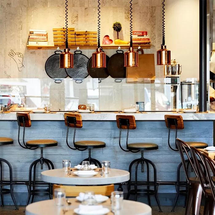 A modern restaurant interior with a marble counter, high wooden stools, hanging copper light fixtures, and shelves with books and decor on a light wall. Round tables are set with plates, glasses, and napkins in the foreground.
