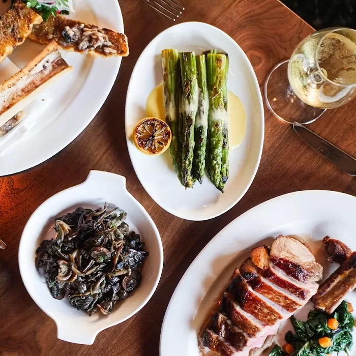 A wooden table set with dishes, including grilled asparagus with sauce, sautéed mushrooms, sliced roasted meat with greens, bone marrow on toast, and a glass of white wine.