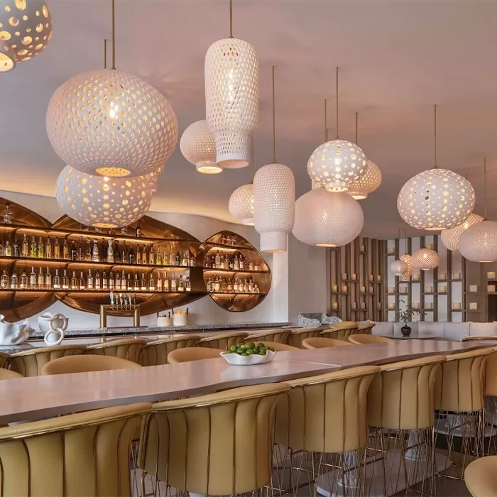 A modern bar with tan leather barstools, a long countertop, decorative light fixtures with intricate designs, and shelves of bottles against a backlit wall with gold oval accents.
