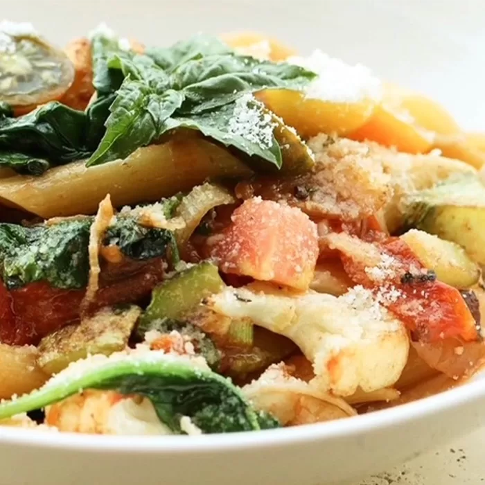 A close-up of a pasta dish with penne, tomatoes, zucchini, leafy greens, grated cheese, and fresh basil, served in a white bowl.