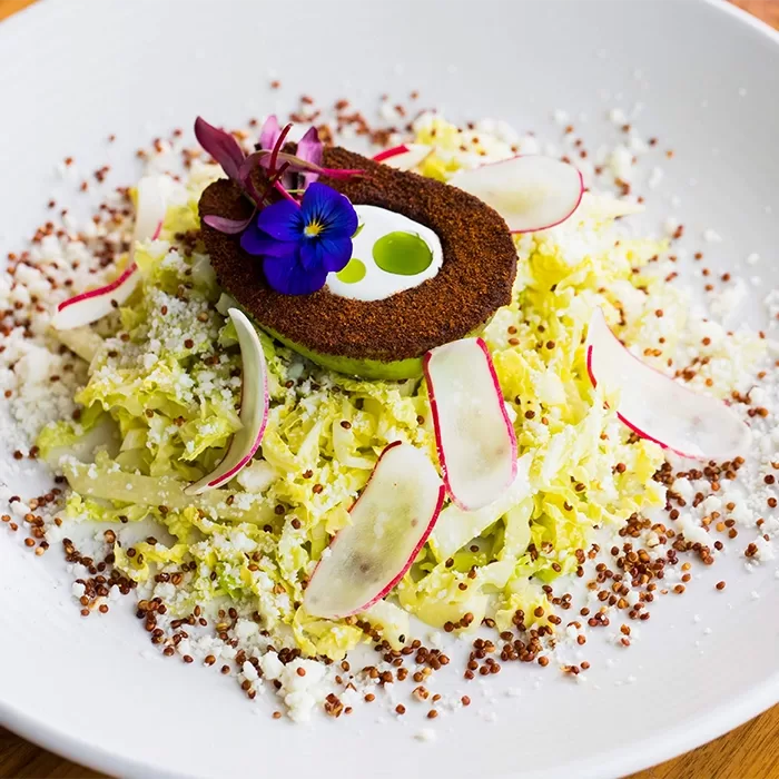 A gourmet salad with shredded cabbage, thin radish slices, red quinoa, crumbled cheese, and a breaded avocado half topped with edible flowers, served on a white plate.