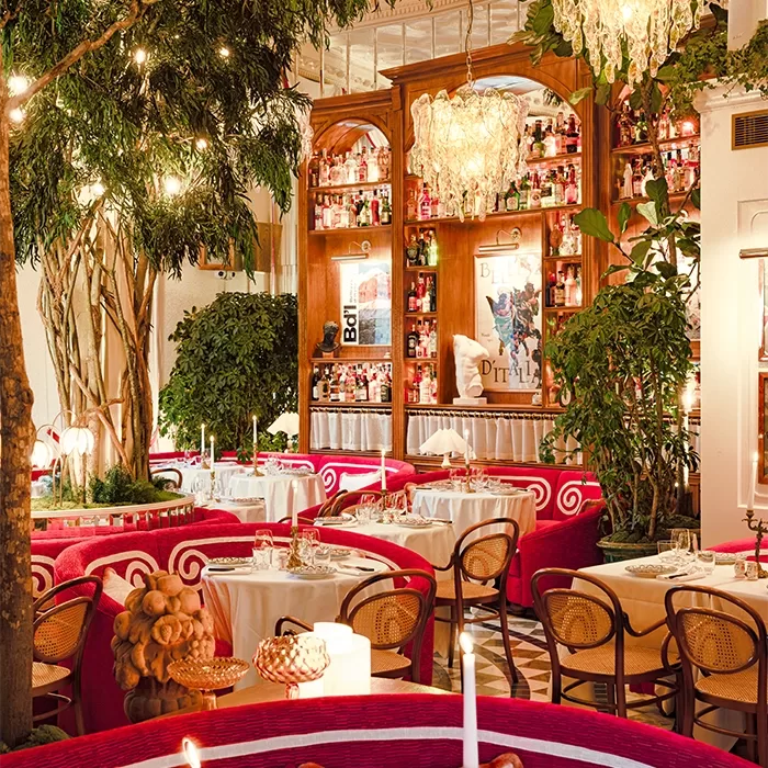Elegant restaurant with red velvet seating, round tables set with white tablecloths, chandeliers, lush indoor plants, warm lighting, and a large wooden bar lined with bottles and decorative mirrors in the background.