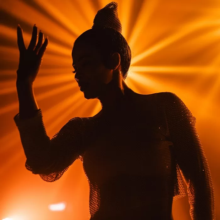 A person poses with one hand raised in front of their face, silhouetted against a dramatic orange backlight that creates radiating beams behind them. The scene feels dynamic and artistic.