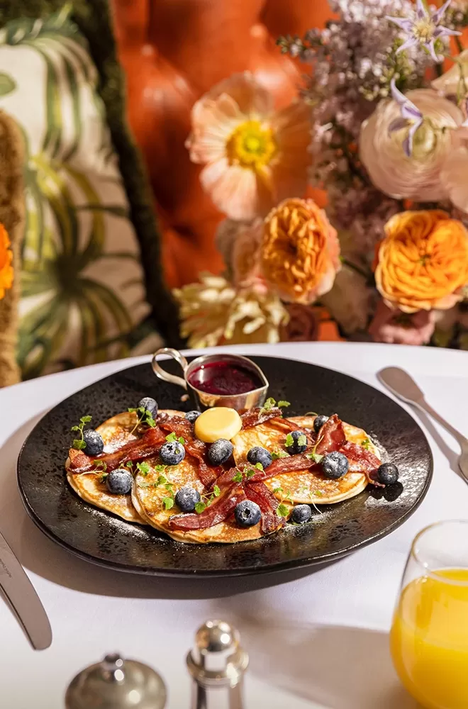 A plate of pancakes topped with blueberries, bacon, and a pat of butter, accompanied by a small jug of syrup. The table is set with a glass of orange juice. In the background, there are vibrant floral decorations at The Ivy, London.