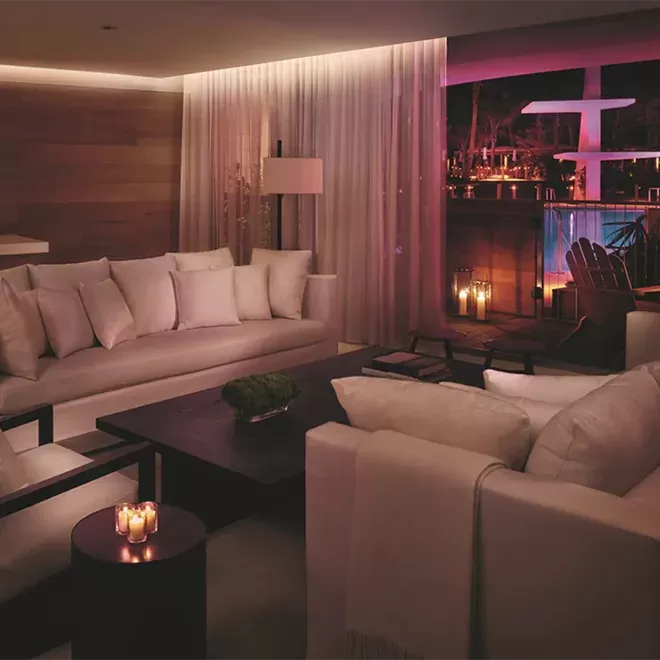 A room at The Miami Beach EDITION hotel beautifully lit lounge with large white couches and a large window.