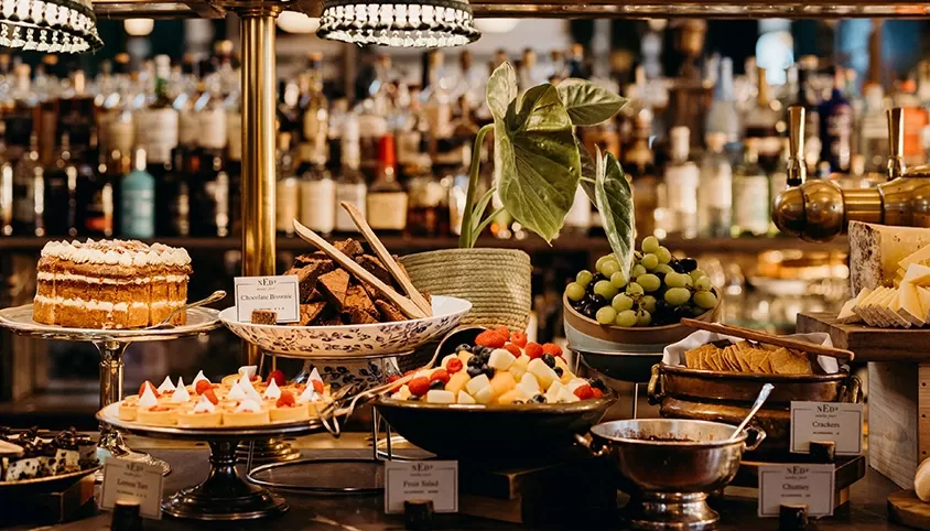 A buffet table with various desserts and appetizers, including a layered cake, fruit skewers, a bowl of grapes, and assorted cheese and crackers. The background displays a well-stocked bar with many bottles at The Ned, London.