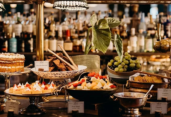 A buffet table with various desserts and appetizers, including a layered cake, fruit skewers, a bowl of grapes, and assorted cheese and crackers. The background displays a well-stocked bar with many bottles at The Ned, London.