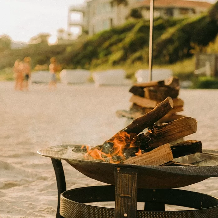 A close-up of a wood fire burning in a metal fire pit on a sandy beach, with blurred people and beach houses in the background.