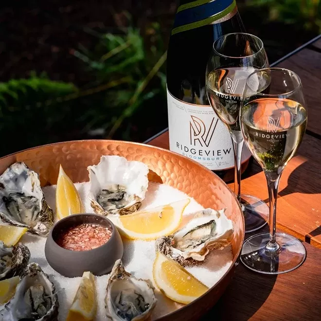 A copper tray holds oysters on a bed of salt with lemon wedges and a small bowl of sauce. Two glasses of white wine and a bottle labeled Ridgeview are beside the tray, set on a wooden table outdoorsat The Rows & Vine at Ridgeview.